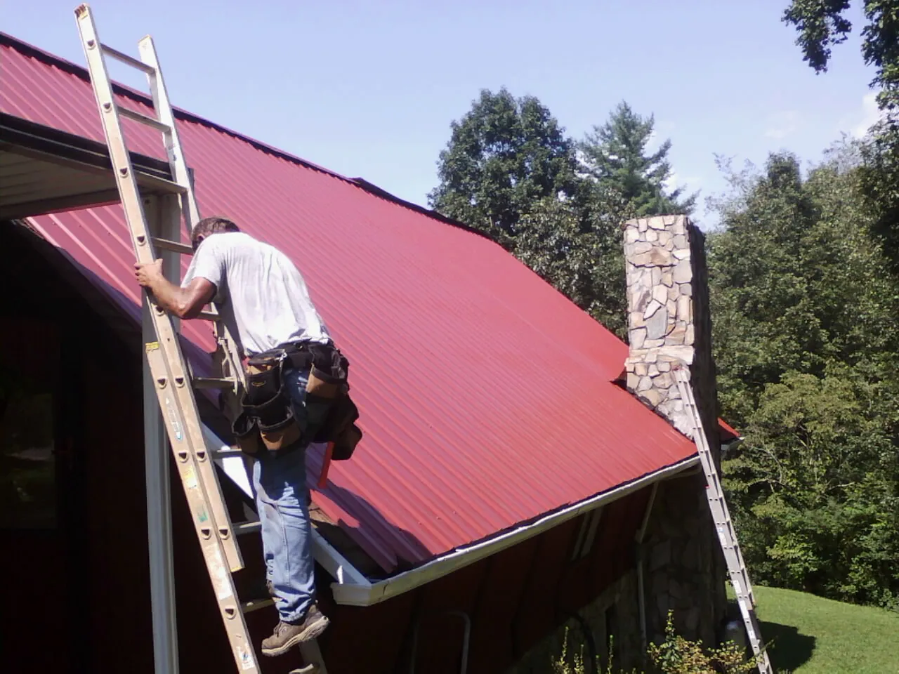 Roofer on a ladder beside a red metal roof and stone chimney — Soffit & Fascia Repair by Roofing Brothers in Tekamah